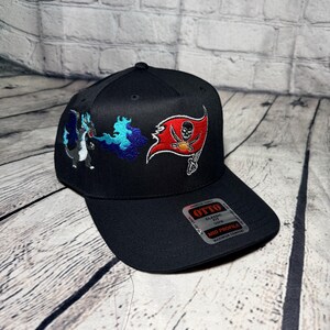 Op de afbeelding: Een zwarte honkbalpet met een geborduurd ontwerp van een blauwe draak die vuur spuugt naast een rode piratenvlag met een schedel en gekruiste beenderen. De pet heeft een platte klep met een Otto-sticker met de tekst Classic Fit, Mid Profile.