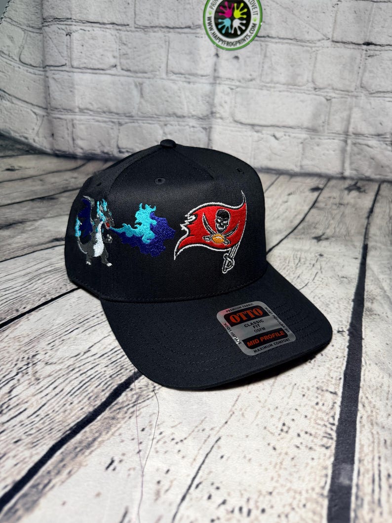 Op de afbeelding: Een zwarte honkbalpet met een geborduurd ontwerp van een blauwe draak die vuur spuugt naast een rode piratenvlag met een schedel en gekruiste beenderen. De pet heeft een platte klep met een Otto-sticker met de tekst Classic Fit, Mid Profile.