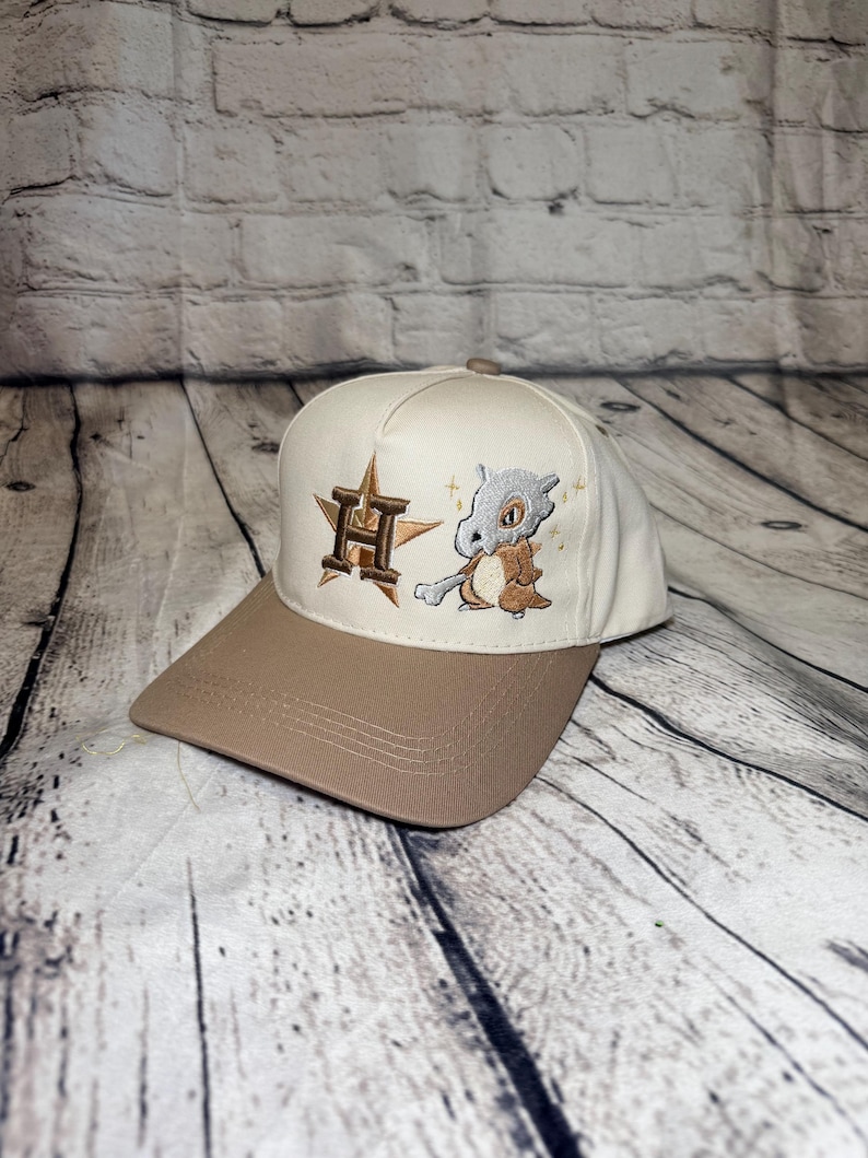 Op de afbeelding: Een cr&egrave;mekleurige baseballpet met een bruine klep. Het voorpaneel is voorzien van een geborduurd bruin en goudkleurig sterlogo met een grote letter H in het midden, naast een geborduurd stripfiguur dat een bot vasthoudt met kleine sterren.