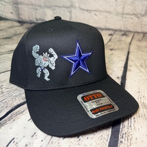 Op de afbeelding: Een zwarte honkbalpet met een geborduurde grijze vierarmige figuur en een blauwe ster. Een sticker op de klep vermeldt: OTTO, CLASSIC FIT, OSFM, MID PROFILE, PREMIUM FABRIC, STYLISH LOOK, MOST POPULAR FIT, MAXIMUM COMFORT.