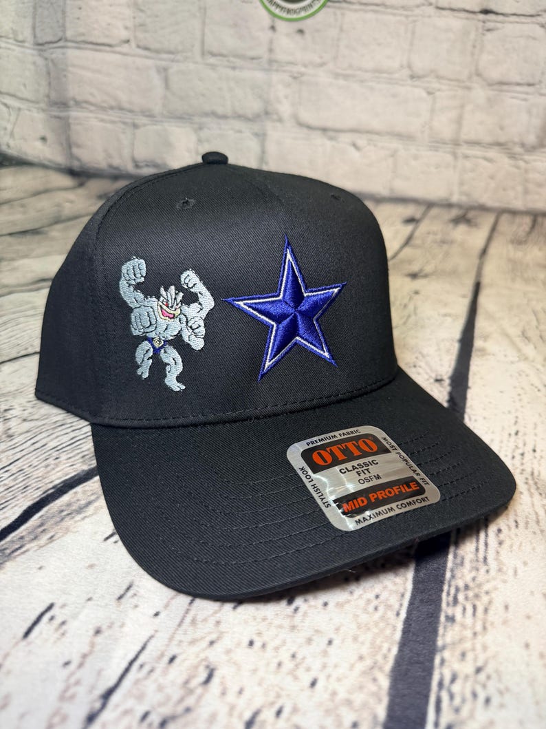 Op de afbeelding: Een zwarte honkbalpet met een geborduurde grijze vierarmige figuur en een blauwe ster. Een sticker op de klep vermeldt: OTTO, CLASSIC FIT, OSFM, MID PROFILE, PREMIUM FABRIC, STYLISH LOOK, MOST POPULAR FIT, MAXIMUM COMFORT.