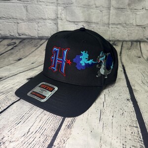 Op de afbeelding: Een zwarte honkbalpet met een geborduurde rode en blauwe letter H, een blauwe wolkachtige vorm en een grijs en blauw draakachtig wezen. Een rechthoekige sticker op de klep toont de tekst OTTO CLASSIC FIT OSFM MID PROFILE.