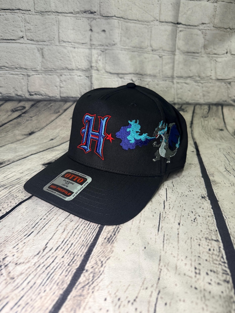 Op de afbeelding: Een zwarte honkbalpet met een geborduurde rode en blauwe letter H, een blauwe wolkachtige vorm en een grijs en blauw draakachtig wezen. Een rechthoekige sticker op de klep toont de tekst OTTO CLASSIC FIT OSFM MID PROFILE.