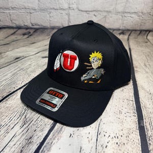 Op de afbeelding: Een zwarte honkbalpet met een geborduurd rood letter U-logo met een witte veer en een geborduurd anime-personage met stekelig geel haar. Een zilveren en zwarte sticker op de klep vermeldt OTTO CLASSIC FIT MID PROFILE.