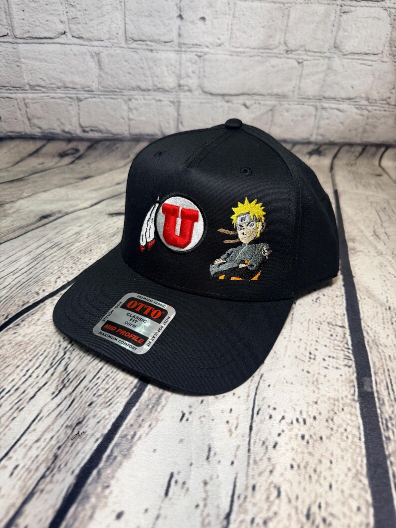 Op de afbeelding: Een zwarte honkbalpet met een geborduurd rood letter U-logo met een witte veer en een geborduurd anime-personage met stekelig geel haar. Een zilveren en zwarte sticker op de klep vermeldt OTTO CLASSIC FIT MID PROFILE.