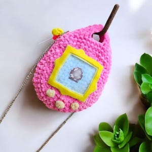 May include: A pink crocheted  handbag shaped like a Tamagotchi, a virtual pet toy. The bag has a yellow screen with a blue background and a silver chain strap.