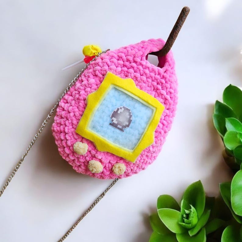 May include: A pink crocheted  handbag shaped like a Tamagotchi, a virtual pet toy. The bag has a yellow screen with a blue background and a silver chain strap.