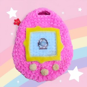 May include: A pink crocheted plush toy shaped like a Tamagotchi, with a yellow frame around a blue screen and three beige pom-poms on the bottom.