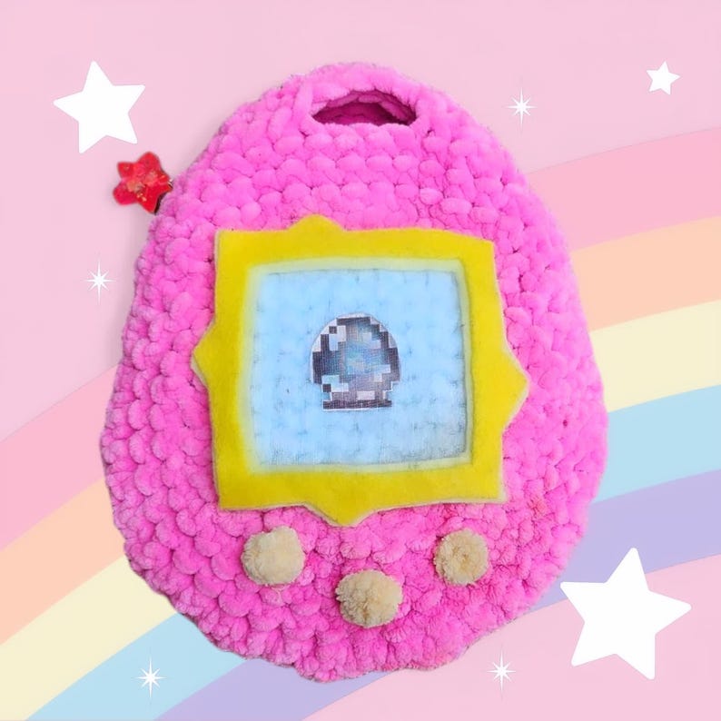 May include: A pink crocheted plush toy shaped like a Tamagotchi, with a yellow frame around a blue screen and three beige pom-poms on the bottom.