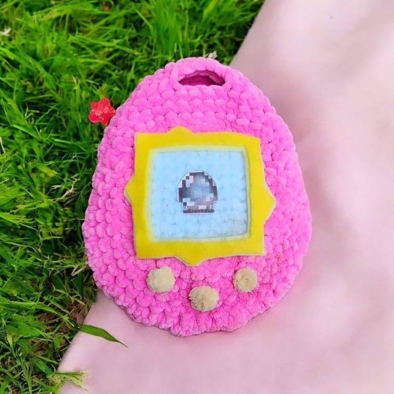 May include: A pink crocheted plush toy shaped like a Tamagotchi with a yellow screen and a blue background. The screen has a pixelated image of a blue and gray object.