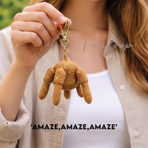 May include: A crocheted brown spider-shaped keychain with yellow-green accents. The keychain is attached to a gold-colored clasp. The text "AMAZE, AMAZE, AMAZE" is at the bottom of the image.