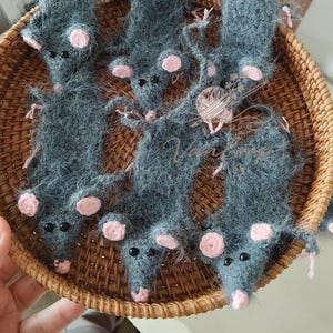 May include: Handmade gray yarn mice with pink ears and noses are arranged in a woven basket. The mice have black bead eyes and long tails. The basket is made of light brown woven material.