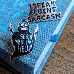 May include: Two enamel pins. One is a silver ghost with the text "See you in hell" and two middle fingers. The other is a silver pin with the text "I speak fluent sarcasm".