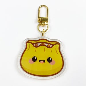 Puede incluir: Un llavero amarillo con forma de dumpling con un cierre dorado. El dumpling tiene una cara sonriente con mejillas rosadas y contornos marrones. El llavero está hecho de acrílico transparente.