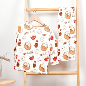 May include: White pajamas with a long-sleeved top and pants featuring a repeated pattern of cartoon dumplings, chopsticks, and soy sauce bottles.