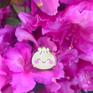 May include: A gold dumpling-shaped pin with a smiling face and pink cheeks, set against a backdrop of bright pink rhododendron flowers. The pin has a cute, cartoon-like design, ideal for adding a touch of fun to any outfit or accessory.