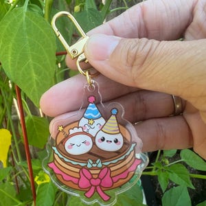 Puede incluir: Un llavero acrílico transparente con un cierre dorado. El llavero presenta una ilustración de dibujos animados de tres personajes sonrientes que llevan sombreros de fiesta sobre una tarta en capas con un lazo rosa.