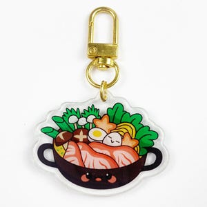May include: A clear acrylic keychain featuring a cartoon illustration of a bowl of ramen. The bowl is brown with a smiling face and contains noodles, meat, vegetables, and an egg. The keychain has a gold-colored clasp.