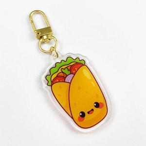 Pode incluir: Um chaveiro de acrílico transparente com um desenho de burrito de desenho animado. O burrito tem um rosto sorridente e é recheado com alface, tomates e cebolas. O chaveiro tem um fecho dourado.