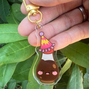 May include: A clear acrylic keychain featuring a cartoon soy sauce bottle wearing a party hat. The bottle is brown with a white top and a smiling face. The keychain has a gold-colored clasp.