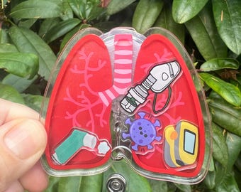 May include: A clear acrylic badge reel featuring a red lung design with medical illustrations. The design includes a nebulizer, a virus, and a pulse oximeter. The badge reel has a clear plastic strap and a metal clip.