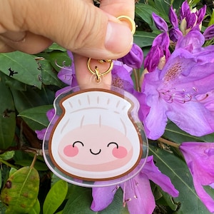 May include: A clear acrylic keychain featuring a smiling cartoon dumpling with pink cheeks and a brown outline. The keychain has a gold-colored ring and clasp. The background includes purple flowers and green foliage.