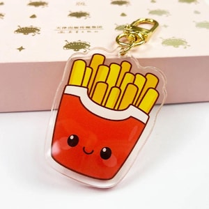 Puede incluir: Un llavero acrílico transparente con una ilustración de dibujos animados de un recipiente de patatas fritas rojo lleno de patatas fritas amarillas. El llavero tiene un cierre dorado.