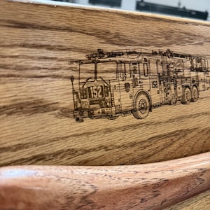 May include: A light brown wooden board featuring a detailed laser-engraved illustration of a fire truck with the number 152 on the front and side. The wood grain is visible throughout the surface, and a curved wooden handle is attached at the bottom.