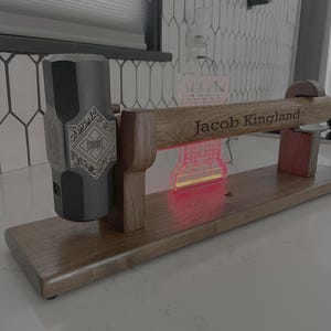 May include: A custom-made wooden hammer display stand with a personalized inscription. The stand holds a large, gray metal hammer with an ornate design on its head. The inscription "Jacob Kingland" is engraved on the wooden handle.