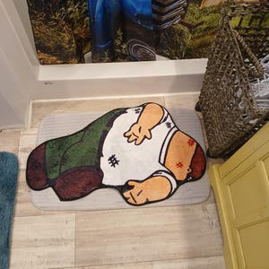 May include: A cartoon-style bath mat featuring a character in a white shirt, green pants, and brown shoes. The mat is on a light gray rectangular rug. The character has red marks on the head and arms.
