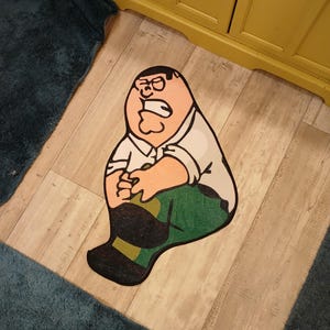Peut inclure: Un tapis de style cartoon repr&eacute;sentant un personnage avec une chemise blanche, un pantalon vert et des chaussures noires. Le tapis a la forme du personnage et est pos&eacute; sur un sol en bois. Le personnage a une expression surprise.