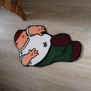 May include: A novelty rug in the shape of a cartoon character, featuring a white shirt, green pants, and a red head. The rug is lying on a light-colored wooden floor.