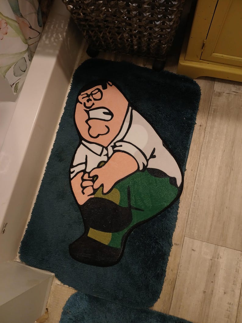 Peut inclure: Un tapis de bain bleu canard repr&eacute;sentant un personnage de dessin anim&eacute; portant une chemise blanche, un pantalon vert et des chaussures noires. Le personnage a une expression surprise. Le tapis est rectangulaire et pos&eacute; sur le sol d'une salle de bain.
