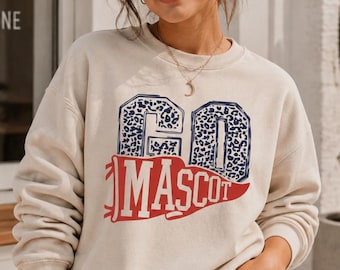May include: A beige crewneck sweatshirt featuring a graphic design with the word GO in a blue leopard print pattern above the word MASCOT in a red pennant shape. The design is centred on the chest of the long-sleeved pullover garment.