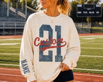 May include: A cream-coloured crewneck sweatshirt featuring a large, distressed navy blue number 21 on the front with the word Custom in red script layered over it. The word NAME is printed in navy blue block letters on the right sleeve.