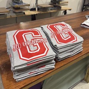 A wooden table with two stacks of gray t-shirts featuring red text and logos, likely representing a school or organization.