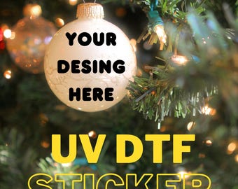 May include: A white Christmas ornament with the text "YOUR DESIGN HERE" in black. The ornament hangs on a Christmas tree with the text "UV DTF STICKER" in yellow. The background is blurred with Christmas lights.