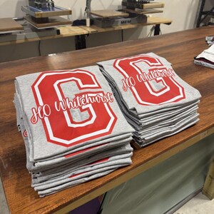 A wooden table with two stacks of gray t-shirts featuring red text and logos, likely representing a school or organization.