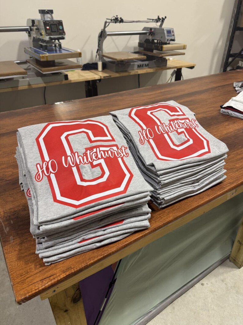 A wooden table with two stacks of gray t-shirts featuring red text and logos, likely representing a school or organization.