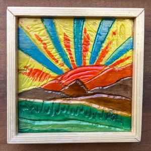 May include: A framed wood carving depicting a sunset over a mountain range. The carving is painted in shades of yellow, orange, red, blue, and green.