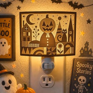 May include: A rectangular plug-in night light featuring a backlit silhouette design of a jack-o'-lantern, a crescent moon, stars, a cat, and a house. The light emits a warm glow from behind the dark cutout panel, which is mounted directly into a standard wall outlet.