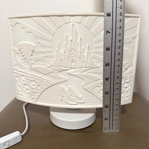 May include: A white, curved, illuminated lamp featuring a relief design of a castle, a winding road, and a pair of shoes. A metal ruler placed vertically next to the lamp indicates a height of approximately 9.75 inches for the decorative panel.