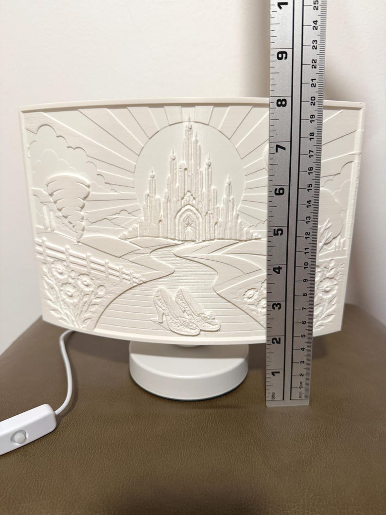 May include: A white, curved, illuminated lamp featuring a relief design of a castle, a winding road, and a pair of shoes. A metal ruler placed vertically next to the lamp indicates a height of approximately 9.75 inches for the decorative panel.