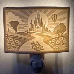 Pode incluir: Uma luz noturna retangular com uma cena de litofania impressa em 3D de um castelo, um tornado, um bal&atilde;o de ar quente e um par de sapatos brilhantes em um caminho de tijolos. A luz brilha por tr&aacute;s, iluminando a paisagem detalhada.