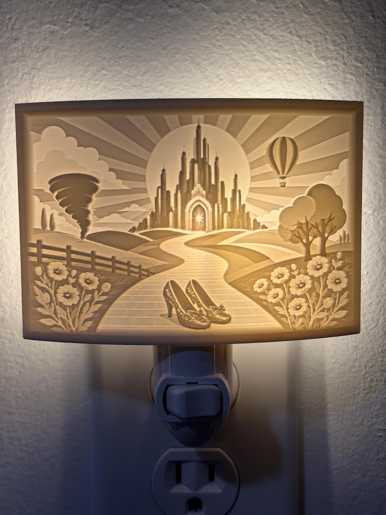 Pode incluir: Uma luz noturna retangular com uma cena de litofania impressa em 3D de um castelo, um tornado, um bal&atilde;o de ar quente e um par de sapatos brilhantes em um caminho de tijolos. A luz brilha por tr&aacute;s, iluminando a paisagem detalhada.