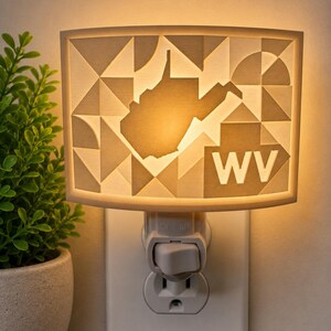 May include: A plug-in night light featuring a rectangular, off-white shade with a geometric pattern of triangles and semi-circles. The center displays a silhouette of the state of West Virginia and the letters WV, illuminated by a warm internal glow.