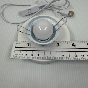 May include: A white circular LED light fixture with a central spherical bulb and two metal spring clips, connected to a white USB power cord with an inline switch. A ruler shows the light fixture has a diameter of approximately 2.5 inches.