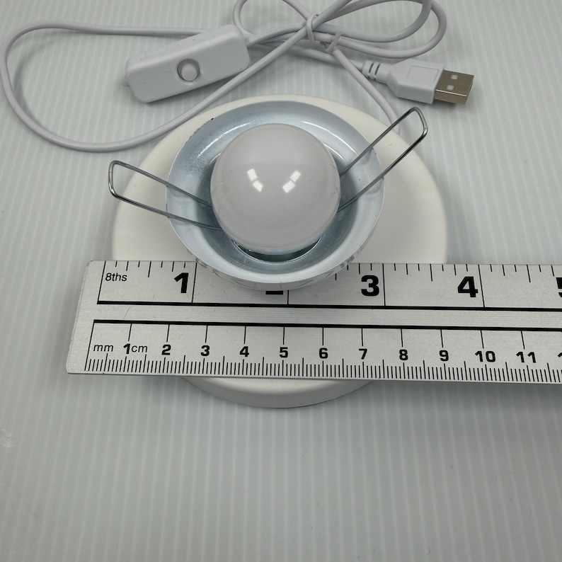 May include: A white circular LED light fixture with a central spherical bulb and two metal spring clips, connected to a white USB power cord with an inline switch. A ruler shows the light fixture has a diameter of approximately 2.5 inches.