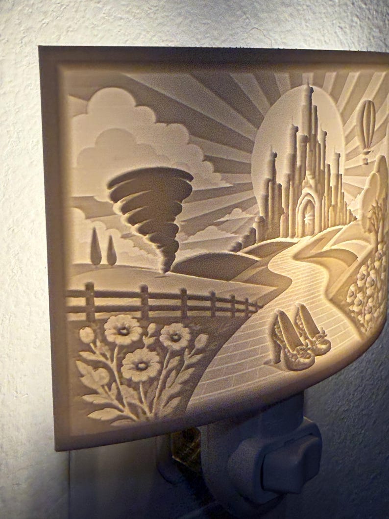 Pode incluir: Uma luz noturna retangular com um relevo de uma paisagem fant&aacute;stica. A cena inclui um tornado, um grande castelo, uma estrada de tijolos sinuosa, um par de sapatos de salto alto, flores em primeiro plano e um bal&atilde;o de ar quente no c&eacute;u.