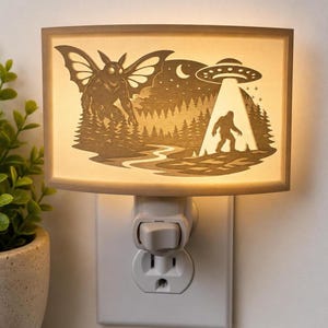 May include: A rectangular plug-in night light featuring a backlit laser-cut design of a moth-like creature, a forest landscape, a crescent moon, and a UFO beaming light down onto a silhouette of a large bipedal figure standing on the ground.